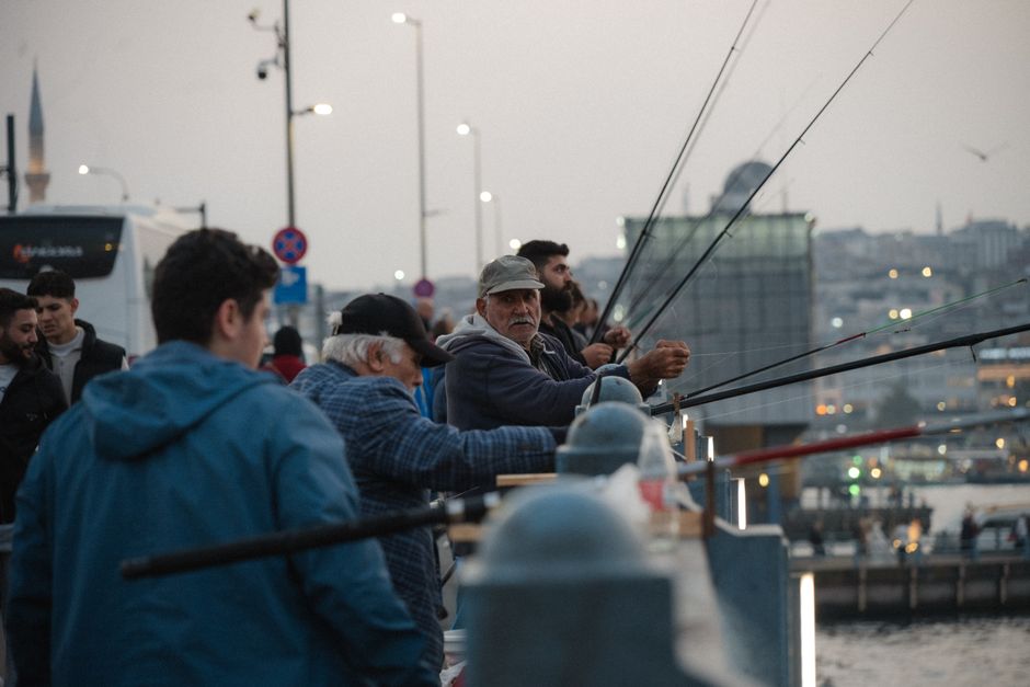A group of men and young people stand along a waterfront promenade, engaged in fishing activities with fishing rods held over the balustrade. In the foreground, a young man in a blue jacket faces away from the camera, while an older man wearing a grey cap and a dark jacket appears to be manually casting or reeling in his fishing line. Other individuals, some with dark beanies or facial hair, are also visible, focused on their fishing. The scene is set during late evening or early dusk, with a grey sky overhead and soft, diffuse lighting creating a calm atmosphere. In the background, city buildings, including a modern glass structure, are visible, along with streetlights and distant boat masts, indicating a busy quay or dock area. The environment suggests a leisurely fishing activity in an urban waterside setting, which could relate to private or independent fishery activities often associated with local or non-official waste and rubbish management around waterfront areas. The image emphasizes natural human interaction with urban outdoor spaces, and the background details hint at a busy port or marina environment, consistent with commercial or recreational fishing activities near a city port.