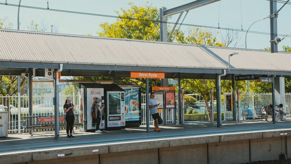 At West Ryde train station, there are four people waiting on the paved platform, which is sheltered by a metal roof supported by grey steel beams. The station sign displaying 'West Ryde' is prominently positioned above the middle of the platform. To the left, a woman wearing a dark shirt, sunglasses, and a hat stands near a bench, looking at her phone. In the center, another woman, also wearing sunglasses and a hat, is interacting with a vending machine or display panel attached to a large, blue advertisement board. To her right, a man in a light-colored shirt, dark shorts, and sunglasses is checking a device, possibly his phone, while standing near an electronic timetable or information display. In the background, there are green trees with yellowing leaves, indicating a possible autumn season, and some utility poles with overhead electrical wires for the train line. Additional signage indicates directions to West Pymble and Hornsby, and there are benches along the platform where a few other individuals are seated. The lighting is natural daylight, casting soft shadows, which amplifies the urban and public transport context. This scene reflects the operational environment of a commuter train station, which may be relevant to private waste disposal services for station or platform cleaning and maintenance, aligning with the context of rubbish management and station waste handling by companies like Rubbish Removal Bayswater.