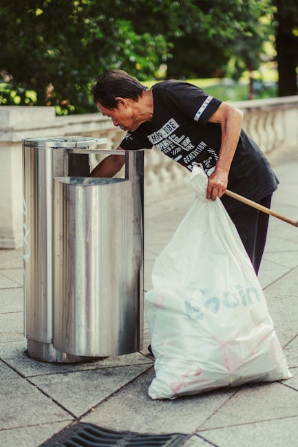 A man with dark hair and light skin is seen disposing of waste into a stainless steel public litter bin located on a paved sidewalk. He is wearing a black t-shirt with partially visible white text, and is bent forward slightly as he places trash into the top opening of the cylindrical bin. In his left hand, he holds a large, semi-transparent white plastic bag filled with rubbish, which appears to contain various waste items. The background shows a low stone or concrete wall and lush green trees, suggesting an outdoor, park-like setting. The scene is lit by natural daylight, emphasizing the reflective surface of the metal bin and the textures of the paving stones. The environment and the presence of the waste disposal unit imply an urban context, likely intended for informal or community waste collection, akin to independent rubbish disposal services like those offered by Rubbish Removal Bayswater. The overall setting conveys responsible waste management, emphasizing proper rubbish disposal in a public space.