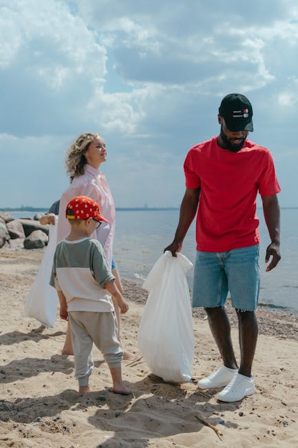 A group of three people, including an adult male, a woman, and a young boy, are participating in a rubbish or waste collection activity on a sandy beach near a body of water. The adult male, wearing a red T-shirt, light blue denim shorts, white sneakers, and a black cap with a logo, is holding a white garbage bag and appears to be gathering rubbish from the sand. The woman, dressed in a light pink jacket and with curly blonde hair, stands nearby observing, while the young boy, wearing a grey T-shirt, light beige shorts, and a red cap with yellow designs, stands with his back to the camera. The background features a cloudy sky, distant shoreline, and large rocks along the water's edge. The scene is outdoors, well-lit by natural daylight, and suggests a community or family effort in responsible waste disposal, aligning with services by Rubbish Removal Bayswater for private waste handling and alternative rubbish collection options.