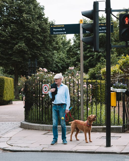A middle-aged man wearing a light blue shirt, dark trousers, and a grey cap stands on a paved sidewalk near a black metal fence, holding a red leash attached to a brown dog with a smooth coat and floppy ears. The man carries a black shoulder bag on his right shoulder and holds a yellow tennis ball in his left hand. Behind him, there is a lush garden with pink and white flowering bushes and green trees, with a sign indicating a 5 miles per hour speed limit positioned near the fence. To the right, an urban street features a black traffic light showing a red pedestrian signal, and a yellow mailbox mounted on a black post is also visible. Suspended above, a blue and yellow directional sign provides street names, including Bayswater and Queensway, indicating the area’s local navigation. The environment appears to be a quiet residential or park-adjacent street during daytime with subdued natural lighting. The scene subtly relates to on-site or private waste disposal options through the context of urban living and pedestrian pathways, while the presence of the dog and signage emphasizes a well-organized neighborhood. Rubbish Removal Bayswater often services such residential and urban environments in the course of regular rubbish collection and waste disposal activities, maintaining the area’s cleanliness and orderliness.