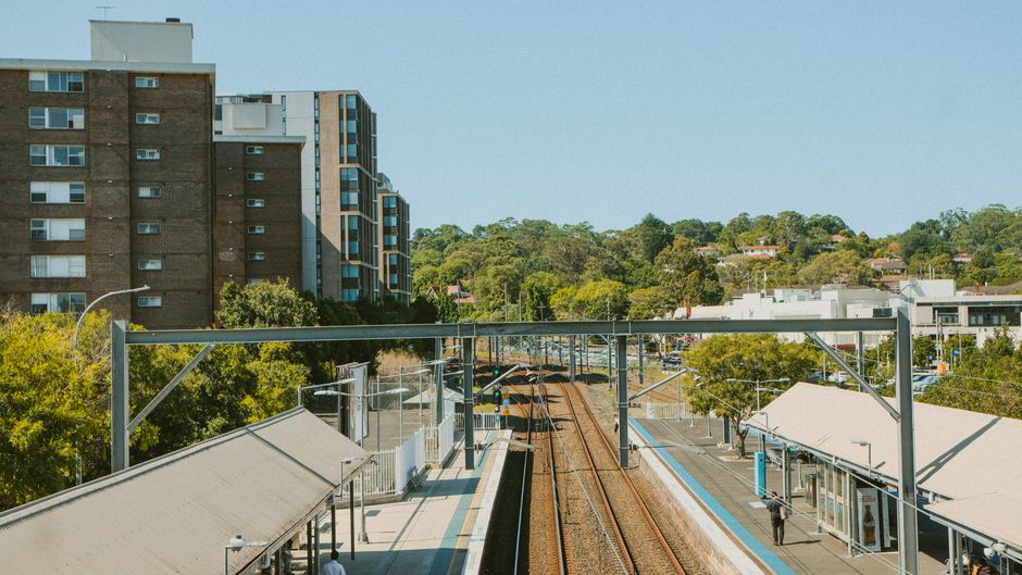 A view of a railway station with two sets of train tracks running through the centre, flanked by platform edges with covered waiting areas and glass barriers. In the foreground, part of the station's roof and the supporting metal framework are visible. On the right side, a person is standing on the platform, possibly waiting for a train. The station is surrounded by trees and residential buildings, including a tall multi-storey apartment block on the left with a brown brick facade and numerous windows. In the background, there are green hills dotted with houses and other structures. The sky is clear and blue, indicating a bright, sunny day. This scene exemplifies an urban transit hub that might be involved in integrated or alternative waste disposal considerations, in line with private rubbish removal services such as those provided by Rubbish Removal Bayswater, which specialize in waste management and clearance at various locations including transport hubs.
