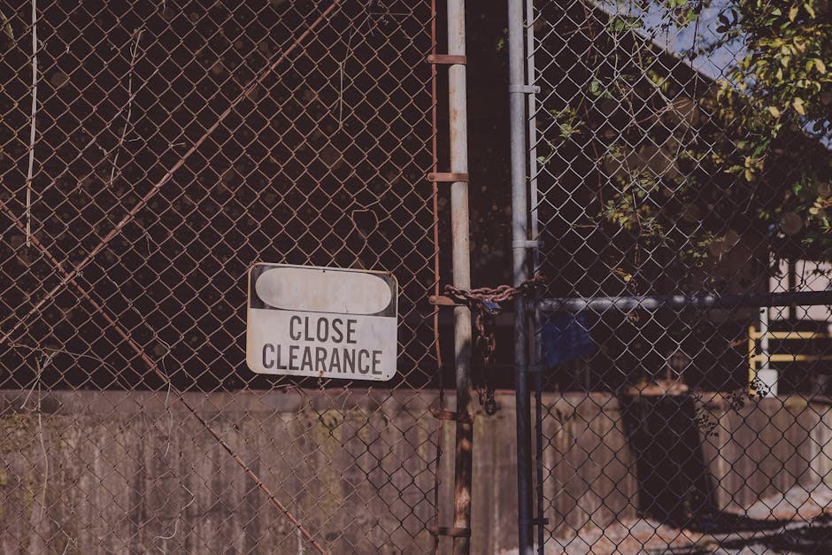 A metal chain and padlock securing a rusted iron gate with a 'Close Clearance' sign attached to the lower section. The gate is part of a chain-link fence that surrounds an outdoor area, possibly a yard or storage space, with dense, dark green foliage visible behind it. The fence posts and gate frame are weathered, with visible rust and corrosion, and the gate hangs slightly ajar on hinges. The surface beneath the fence appears to be concrete or pavement, with some dirt and moss at the base. Sunlight illuminates the scene, casting shadows on the fence and highlighting the textured, weathered surfaces, suggesting it is an outdoor setting for private or restricted access, relevant to waste management or rubbish removal services operating in secure environments. Rubbish Removal Bayswater may handle similar secured clearance areas for waste collection or disposal activities, emphasizing the importance of controlled access in private waste procedures.