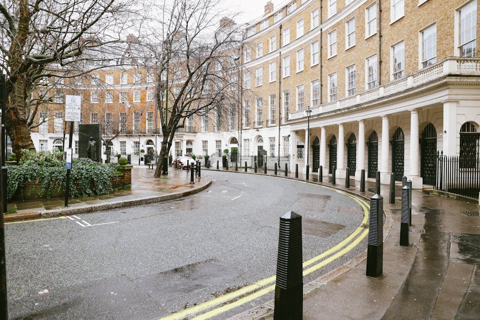 A curved residential street scene with a wet asphalt road and double yellow lines along the curb. On the right side, there is a tall white Victorian-style building with black iron gates and black bollards spaced evenly along the pavement, serving as parking restrictions or barriers. Several leafless trees with thin branches are visible in the background, alongside a small square area with a statue or monument on a pedestal surrounded by low, neatly trimmed bushes. The building facades are composed of light-colored brick with large, uniform sash windows, some of which reflect the overcast sky and surrounding environment. The overall scene appears during a rainy day, evident from the damp pavement and subdued natural lighting, and it is situated in an urban setting where private waste collection or rubbish removal services might be used as part of property maintenance and clearance activities, consistent with the context of rubbish removal in a residential area.