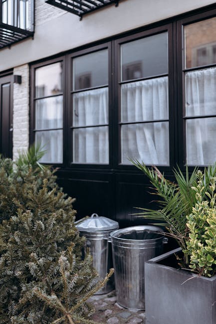 The image shows an exterior scene of a residential building with a dark-colored bay window featuring multiple glass panes divided by slim black frames. The window is partially covered by white curtains visible through the glass. In front of the window, there is a small arrangement of outdoor potted plants, including a bushy green shrub with dense, needle-like leaves and another plant with long, slender, spiky leaves. Positioned on the paved ground near the windows are three metallic rubbish bins with rounded lids and smooth, reflective surfaces. The bins are placed close together, with one partially hidden behind the shrub and another slightly in front. The background includes a brick wall and the ground appears to be grey paving stones, suggesting an outdoor patio or garden area. Overall, the scene conveys a tidy outdoor space that may be used for domestic waste disposal or collection, with a subtle emphasis on private rubbish management services that Rubbish Removal Bayswater offers, especially suited for properties with limited access. The natural lighting is neutral, with no strong shadows, indicating an overcast day or shaded environment.