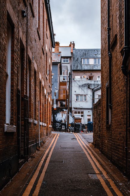 A narrow alleyway between two multi-storey brick buildings with a slightly wet pavement marked by two yellow lines on each side, leading towards a residential area with various structures including a wooden staircase, a grey concrete building with small windows, and a garage door in the background. The alley is enclosed by brick walls on both sides, with visible black drainpipes and window frames. The scene appears overcast with diffuse lighting, and the surrounding environment suggests an urban setting where private waste collection or rubbish removal may be necessary, aligning with local waste management practices. Rubbish Removal Bayswater may occasionally provide services in similar narrow access situations, offering tailored waste clearance options for such confined spaces.